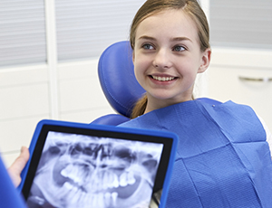 Young girl in dental chair smiling as dentist reviews her digital x-rays